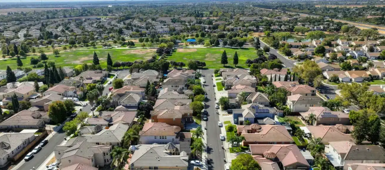Aerial view of a suburban neighborhood with neatly arranged houses, green parks, and tree-lined streets under a clear blue sky.