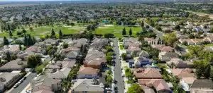 Aerial view of a suburban neighborhood with neatly arranged houses, green parks, and tree-lined streets under a clear blue sky.